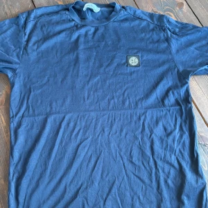 Blå t-shirt från Stone Island - Snygg marinblå t-shirt från Stone Island med klassisk logopatch på bröstet. T-shirten har rund hals och korta ärmar, tillverkad i mjuk bomull som känns skön mot huden. Mycket bra skick. Perfekt för dig som gillar stilrena och ikoniska plagg.