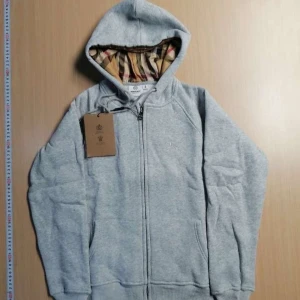 Grå hoodie från Burberry med dragkedja - Snygg grå hoodie från Burberry med klassiskt rutigt foder i huvan. Tröjan har dragkedja framtill, snörning i huvan och två fickor. Perfekt för dig som gillar stilrena plagg med en touch av lyx. Det är Xs 
