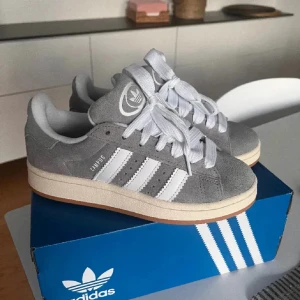 Grå Adidas Campus sneakers - Snygga grå Adidas Campus sneakers i mocka med vita klassiska ränder och vit sula. Skorna har rund tå, platt sula och snörning. Perfekta för dig som gillar en clean och tidlös stil. Använda fåtal gånger men är som nya i skicket. 