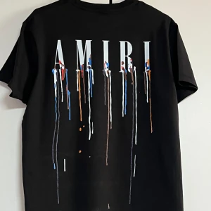 Svart AMIRI t-shirt med färgstänk - Svart t-shirt från AMIRI med stort tryck på ryggen och mindre tryck fram, båda med vita bokstäver och färgstänk i blått, rött, orange och vitt. Klassisk passform och rund hals. Perfekt för dig som gillar streetstyle och statement-plagg.