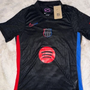 Barcelona x Nike fotbollströja  - Snygg svart FC Barcelona matchtröja från Nike med klubbmärke på bröstet, röd och blå detalj på sidorna och röd Spotify-logga framtill. Tröjan har korta ärmar, V-ringning och är tillverkad i lätt, ventilerande funktionsmaterial. Passar alla årstider och är stilig året om. 