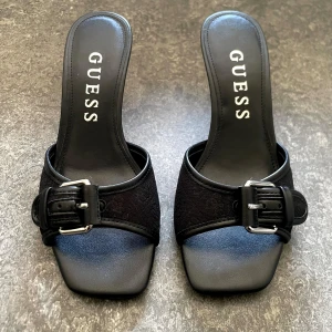 Svarta klackade sandaler från Guess - Stilrena svarta klackar från Guess med öppen tå  och smyckat silver spänne. Klackarna är tillverkade i skinn med diskret mönster på remmen. Perfekta för dig som vill ha en trendig och elegant look.