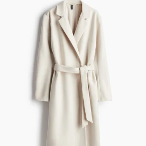 Beige lång kappa med bälte från H&M - Stilren beige kappa från H&M med klassisk krage och knytskärp i midjan. Lång modell med raka ärmar och snyggt fall. Perfekt för dig som vill ha en clean och tidlös look. Passar lika bra till jeans som till klänning.