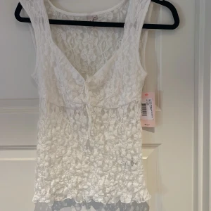 Vit spetsblus från Nelly - Romantisk vit blus från Nelly i genombruten spets med v-ringning och knyt framtill. Ärmlös modell med fina spetsdetaljer och lätt transparent look. Perfekt för dig som vill ha en feminin och trendig topp med extra detaljer.