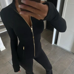 Svart träningsjacka från Lululemon - Snygg svart träningsjacka från Lululemon med guldiga dragkedjor och figurnära passform. Jackan har hög krage, långa ärmar och två fickor med dragkedja. Perfekt för dig som vill ha en stilren look till gymmet eller på språng.