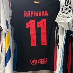 FC Barcelona x Nike Cactus Jack Travis Scott fotbollströja Raphinha - Snygg svart FC Barcelona fotbollströja i samarbete med Cactus Jack och Nike. Tröjan har röda detaljer, tryck med Raphinha 11 på ryggen, samt UNHCR-logga. Framsidan har ett svart FCB-märke med blå och röd kant, samt unikt graffiti-tryck. Tillverkad i lätt och ventilerande material.