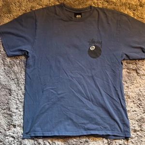 Blå Stüssy t-shirt med 8-ball print - Blå t-shirt från Stüssy med stort 8-ball tryck och logga på ryggen. Slight wear på printen på ryggen