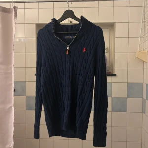 Marinblå kabelstickad tröja Polo Ralph Lauren - Kabelstickad marinblå tröja från Polo Ralph Lauren med half zip och röd logga Strl l