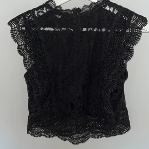 Svart spetsblus med volangdetaljer - Supersnygg svart blus i spets med fina volangdetaljer längs ärmar och nederkant. Hög halsringning och ärmlös design ger en trendig look. Perfekt att styla med jeans eller kjol för en cool outfit.