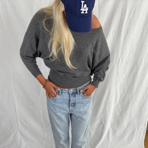 Mörkgrå offshoulder stickad tröja - Snygg mörkgrå stickad tröja med offshoulder-look och bred mudd i midjan. Tröjan har lång ärm och en loose passform som ger en avslappnad vibe. Perfekt att styla med jeans för en chill och trendig outfit.