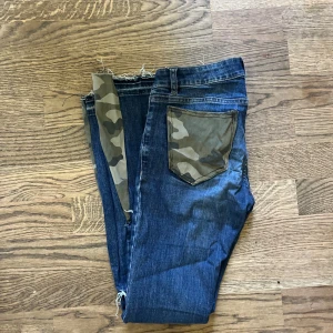 Bootcut jeans med camo-detaljer - Unika blå jeans från Lindex med bootcut passform, camouflage-tyg på ena bakfickan och längs bensluten. Snygga slitningar på benen och råa kanter ger en cool vibe. Perfekta för dig som vill sticka ut med detaljer och mönster.