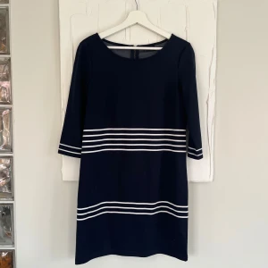 Marinblå kortklänning med vita ränder - Snygg marinblå klänning med vita horisontella ränder och trekvartsärm. Klänningen har rund halsringning och dragkedja bak. Fint skick 🥰