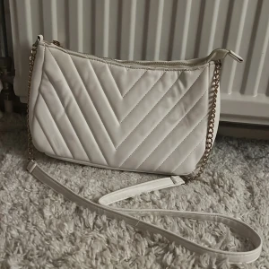 Vit quiltad axelväska med kedja - Snygg vit axelväska med quiltat mönster i V-form och guldfärgad kedjedetalj på axelremmen. Väskan har dragkedja upptill och ett rymligt innerfack med extra ficka. Perfekt storlek för det viktigaste och ger en clean look till din outfit.