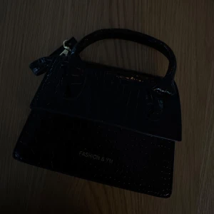 Svart handväska  - Stilren svart handväska med croco-mönstrad yta och kort handtag. Väskan har en klassisk, fyrkantig form och detaljer i guld. Perfekt för dig som vill ha en trendig accessoar med lyxig känsla.