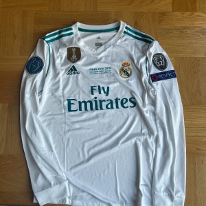 Real Madrid Ronaldo 7 Finaltröja 2018 - Real Madrid långärmad fotbollströja från Champions League-finalen 2018 i Kiev, med Ronaldo 7 på ryggen. Vit med turkosa detaljer, broderat klubbmärke, FIFA-märke och Champions League-patchar. Tillverkad i lätt och ventilerande polyester (Climacool).