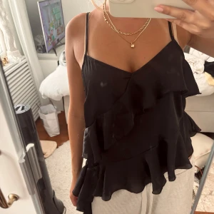 Svart volangig linnetopp Gina Tricot - Supersnygg svart linnetopp från Gina Tricot med tunna axelband och flera lager volanger. Toppen har v-ringning och asymmetrisk nederkant. Perfekt festtopp.