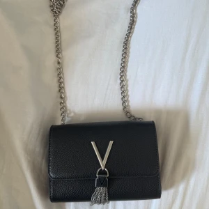Svart axelväska från Valentino - Snygg svart axelväska från Valentino med silverfärgad kedja och ikoniskt V-spänne framtill. Väskan är i skinn med struktur och har dekorativa silverdetaljer och fransar. Perfekt storlek för det viktigaste och ger en lyxig vibe till din outfit. Använd tre gånger. Original pris 500kr