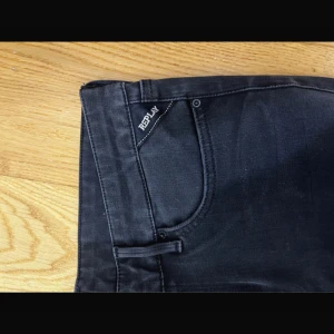 Svarta Replay anbass jeans - Säljer ett par svarta jeans från Replay med klassisk femficksdesign och diskret logga vid fickan. Jeansen har smal passform och raka ben, tillverkade i mjukt denimtyg som sitter snyggt. Perfekta för dig som gillar stilrena och bekväma jeans.
