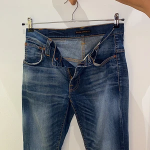 Nudie Jeans blå slim fit jeans - Snygga blå jeans från Nudie Jeans med klassisk orange söm och cool slitning. Jeansen är tillverkade i 100% ekologisk bomull och har en slim passform med raka ben. Bakfickorna har Nudies ikoniska broderi. Perfekta för en avslappnad streetstil.