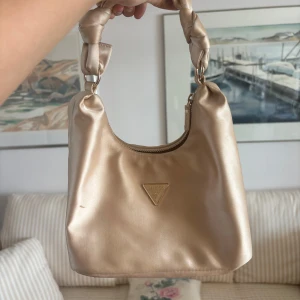 Champagnefärgad axelväska från Guess - Snygg axelväska från Guess i champagnefärgat satinliknande material. Väskan har ett knutet handtag, dragkedja upptill och den klassiska Guess-loggan i metall framtill. Perfekt storlek för det viktigaste och ger en lyxig touch till din outfit. OBS se defekter i bilderna 🩷