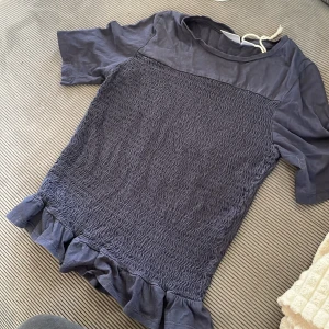 Mörkblå topp med volang från Gina Tricot - Supersöt mörkblå topp från Gina Tricot med korta ärmar och smockad framsida. Toppen har en volangkant nedtill som ger en extra touch. Perfekt för dig som gillar en lite sötare stil och vill ha något som sticker ut.