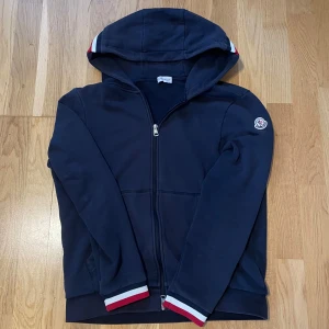 Mörkblå Moncler hoodie/kofta med huva - Snygg mörkblå hoodie från Moncler med dragkedja, huva och långa ärmar. Ribbade muddar med röd, vit och blå rand. Moncler-logga på ärmen och klassisk design. Perfekt för dig som gillar stilrena och exklusiva plagg. Den är i storlek L men sitter ungefär samma som en M. Skriv gärna vid frågor/fler bilder.