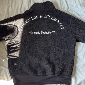 Svart teddyjacka från ASOS Design - Mysig svart teddyjacka från ASOS Design med hög krage och snörning framtill. Baksidan har vit brodyr med texten 'Forever & Eternity' och 'Dark Future'. Perfekt för kalla dagar och ger en cool streetstyle-vibe. Passar storlekarna S, M och L beroende på önskad passform. Nypris ca 500kr.