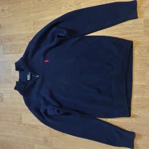 Mörkblå stickad tröja Polo Ralph Lauren - Mörkblå stickad tröja från Polo Ralph Lauren med klassisk röd broderad logga på bröstet. Tröjan har ribbade muddar vid ärmslut och nederkant samt långärmad passform. Perfekt för dig som gillar stilren och tidlös design.