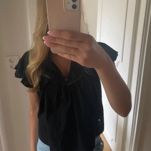 Svart blus med volangärm - Svart blus med v-ringning och volangärmar. Lätt och luftig modell med lös passform, perfekt till jeans eller kjol. Tunt tyg med diskret spetsdetalj längs bröstet. Enkel och stilren, passar dig som gillar en avslappnad men trendig look. Väldigt bra skick 😊