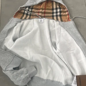 Grå Burberry hoodie - Snygg grå hoodie från Burberry med ikoniskt rutigt foder i huvan. Tröjan har dragkedja, långa ärmar och ribbade muddar. Tillverkad i mjuk bomullsmix och har snörning i huvan för extra stilpoäng. Perfekt för dig som gillar klassisk design med lyxig touch.