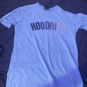 Blå HOODRICH t-shirt XS - Snygg blå t-shirt från HOODRICH i storlek XS. T-shirten har korta ärmar och ett stort broderat HOODRICH-tryck i olika nyanser framtill. Skön bomullskvalitet och klassisk passform, perfekt för en chill streetwear-look.