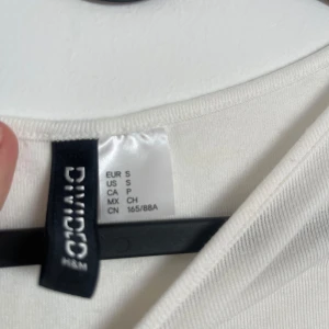 Vit T-shirt från H&M Divided - Aldrig använd T-shirt med rynkningar på sidorna som gör tröjan till en jättefin modell. Köpt på h&m i storlek s men den har väldigt stretchiga material och är i en väldigt fin kröning vit färg.