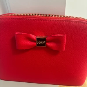 Röd axelväska Holly & Whyte med rosett - Snygg röd axelväska från Holly & Whyte med guldfärgad kedja och dragkedja. Framsidan har en söt rosett med H&W-detalj. Väskan är i slätt syntetmaterial och har ett innerfack med dragkedja. Perfekt för att lyfta din outfit med en färgklick!