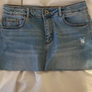 Ljusblå jeanskjol med slitning - Snygg ljusblå jeanskjol med rå kant och slitning på ena sidan. Klassisk femficksmodell med knapp och dragkedja framtill. Perfekt för dig som gillar en avslappnad och trendig stil.