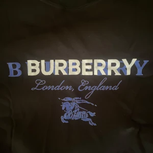Svart Burberry sweatshirt - Svart sweatshirt från Burberry med blått och vitt tryck på bröstet där det står 'Burberry London, England' och en riddare. Tröjan har rund hals och långa ärmar, perfekt för en stilren och chill look.