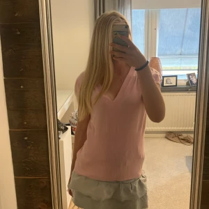 Ljusrosa v-ringad topp från Gianini - Supersöt ljusrosa topp från Gianini med v-ringning och korta puffärmar. Toppen har ribbad nederdel som ger en snygg passform och är tillverkad i ett mjukt, stretchigt material. Perfekt att styla med kjol eller jeans för en fräsch look.