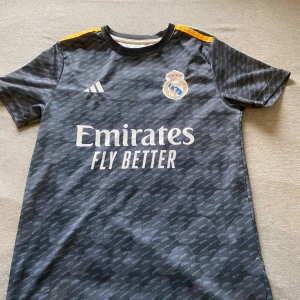 Real Madrid Bellingham tröja Adidas 164 - Snygg Real Madrid fotbollströja från Adidas med Bellingham och nummer 5 på ryggen. Tröjan är svart med subtilt mönster, gula detaljer på axlarna och lila inslag. Tillverkad i lätt och ventilerande ClimaCool-material. Perfekt för dig som älskar fotboll och Real Madrid.