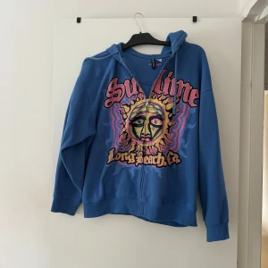 Blå Sublime hoodie från H&M - Säljer en blå hoodie från H&M med Sublime-tryck och färgglad grafik på framsidan. Tröjan har huva, dragkedja och långa ärmar. 