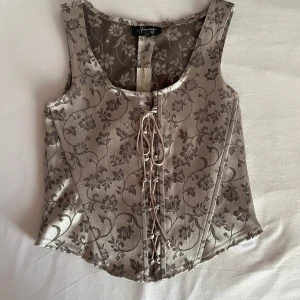 Blommig silvrig väst/korsett med snörning - Supersnygg väst i silvrig nyans med blommigt mönster och snörning framtill. Västen har en silkeslyster och är figursydd med breda axelband. Perfekt för att lyfta vilken outfit som helst med en touch av vintagekänsla. Aldrig använd 