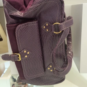 Lila axelväska i ormskinnsmönster - Säljer en snygg lila axelväska med ormskinnsmönster och detaljer i mocka. Väskan har guldiga spännen och ringar, samt en stor ficka framtill med bälte. Om man vill kan köpa stora väskan + de två mindre tillhörande. Eller separat. 