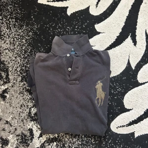 Polo ralph lauren Big pony tröja - Svart långärmad pikétröja från Polo Ralph Lauren i custom fit. Tröjan har klassisk krage, knäppning med två knappar och stort broderat polospelare-logo på bröstet samt en guldfärgad siffra 3 på ärmen. Tillverkad i mjuk bomull.