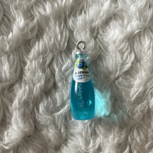 Blåbärsflaska berlock. - Supergulligt berlock format som en blåbärsdricka i miniatyr. Hängsmycket är blått med glittereffekt och har en etikett med blåbärsmotiv. Perfekt för dig som gillar unika och lekfulla accessoarer.
