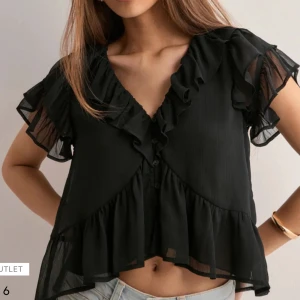 Svart volangblus med v-ringning - Supersöt svart blus med v-ringning och volangdetaljer längs halsringningen och ärmarna. Blusen är croppad och har knappar framtill. Aldrig använd endast testad! Storlek 34 passar XS-S