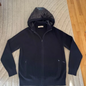 Svart stickad jacka med quiltad huva - Svart stickad jacka från SayLess med quiltad, glansig huva i syntetmaterial. Jackan har dragkedja framtill, två fickor med dragkedja och ribbade muddar. Perfekt för dig som gillar en clean och stilren look med lite edge.