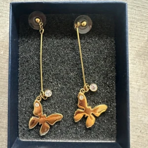 Guldiga örhängen med fjärilar - Snygga långa örhängen i guld med hängande fjärilar och små glittrande stenar. Perfekta för dig som vill sticka ut med en cool och trendig accessoar. Fjärilarna ger en lekfull vibe och de gnistrande detaljerna gör dem extra speciella.