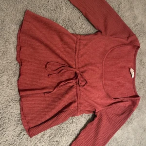 Ribbad röd topp med knyt i midjan - Supersnygg ribbad röd topp med lång ärm och knyt i midjan. Den har en rundad urringning och är tillverkad i ett mjukt, stretchigt material som sitter bekvämt. Perfekt för dig som gillar en feminin och trendig stil.