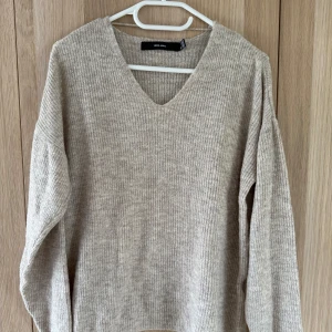 Beige ribbad stickad tröja Vero Moda - Supermjuk beige stickad tröja från Vero Moda med v-ringning och ribbad struktur. Långärmad och i en klassisk, lite loose passform som är perfekt för lager-på-lager. Enkel att matcha med jeans eller kjol för en chill look.