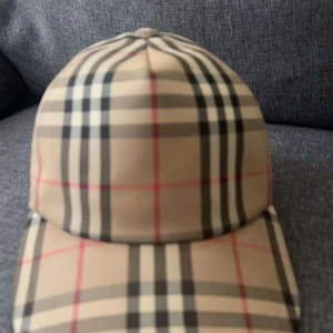 Rutig keps från Burberry - Snygg keps från Burberry i klassiskt beige rutigt mönster med svarta, vita och röda linjer. Justerbar baktill med svart spänne och tydlig Burberry-logga. Perfekt accessoar för en trendig streetstyle-look.
