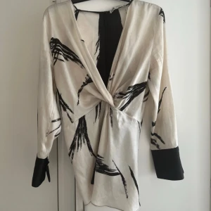 Vit och svart omlottklänning/blus med knyt - Snygg kortklänning/blus i vit och svart med abstrakt mönster. Omlott framtill med twist-detalj i midjan, långa ärmar med svarta muddar och guldknappar. Materialet känns som satin eller polyester, perfekt för dig som gillar unika detaljer. Saknas en guldknapp på ena ärmen. 