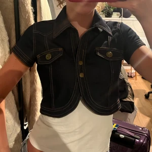 Jeans bolero korta ärmar - Mörkblå bolero från River Flower med korta ärmar, markerade sömmar och två bröstfickor med knappar. 